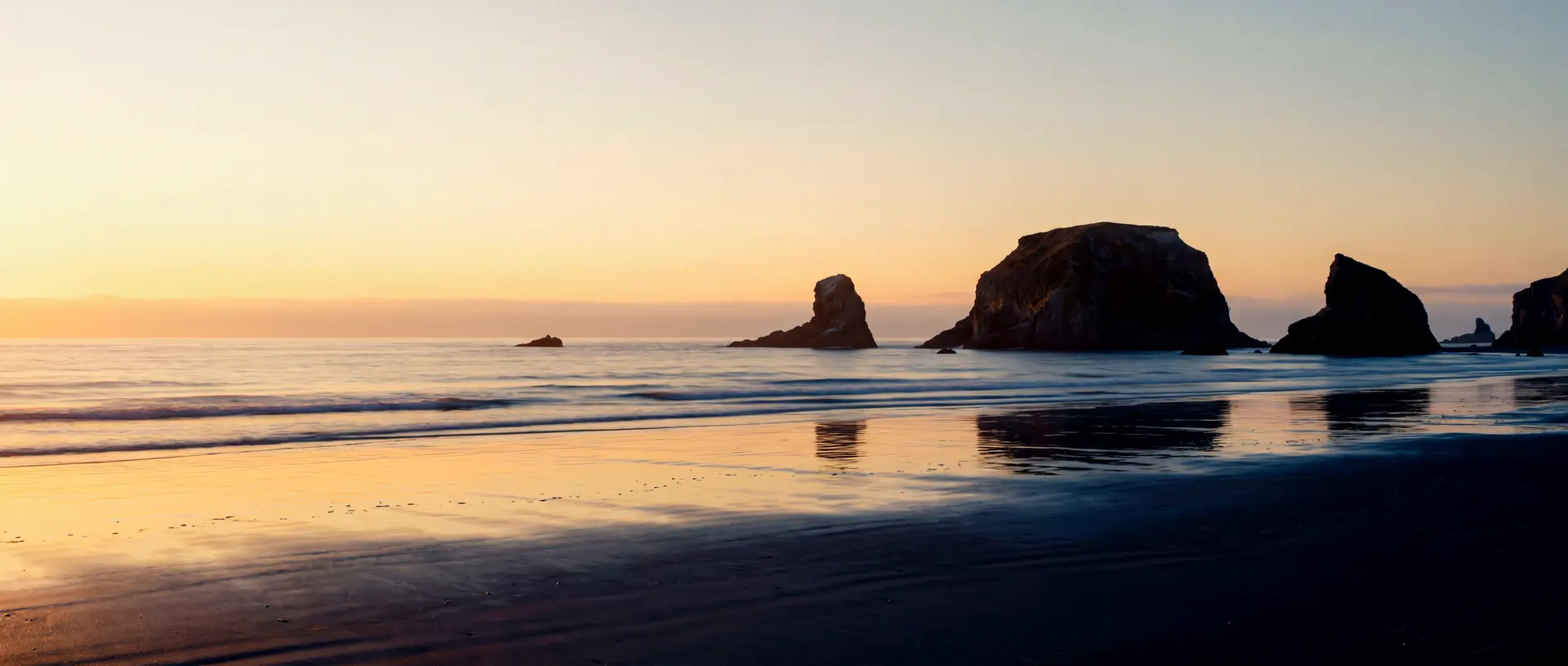 The Bandon, Oregon coastline at golden hour, sea stacks standing in a calm Pacific with wet sand reflecting the warm sky.