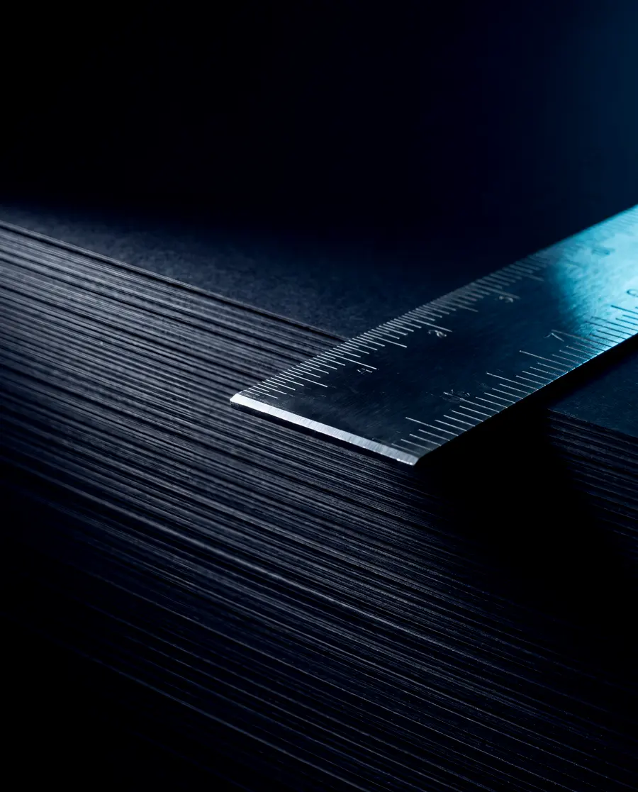A machinist's steel rule laid along the edge of a thick stack of matte black cards, cyan light raking across the metal.