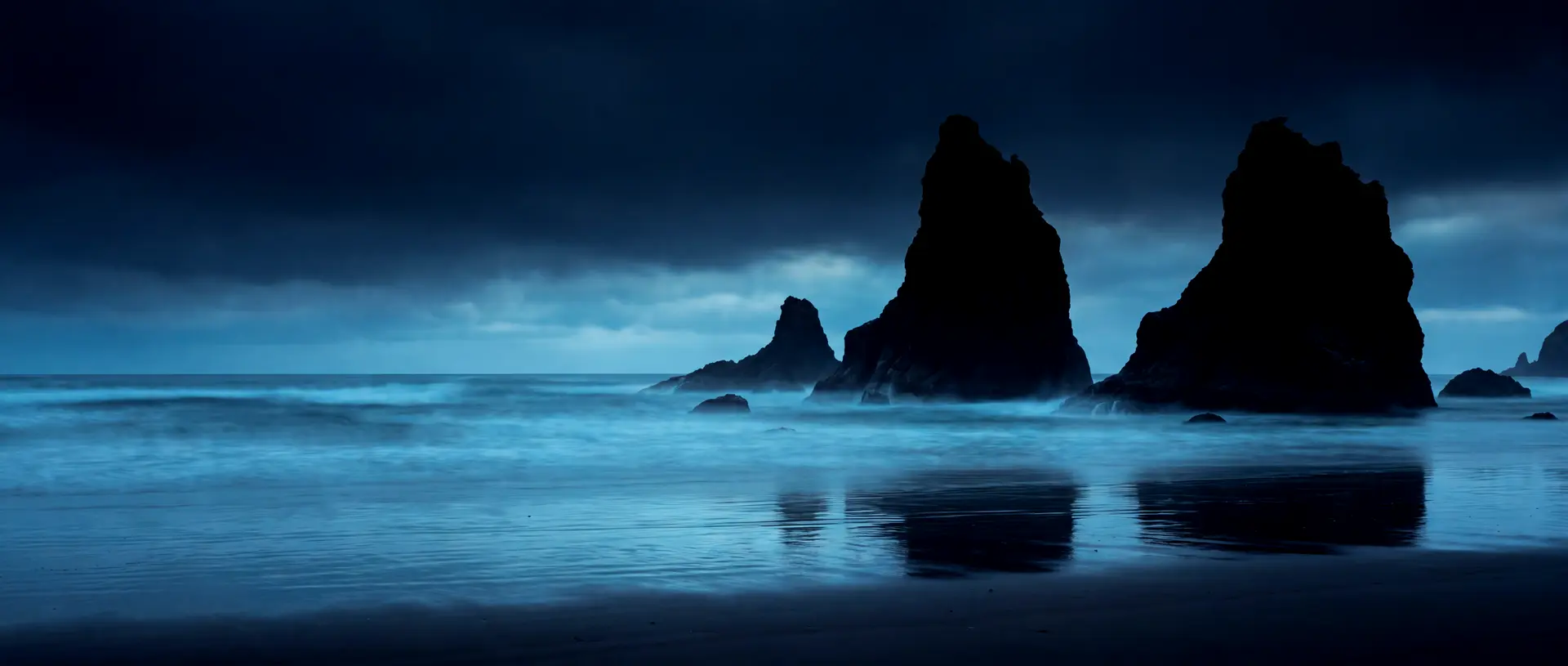 Basalt sea stacks off the southern Oregon coast at blue hour, the surf gone to mist around their bases.